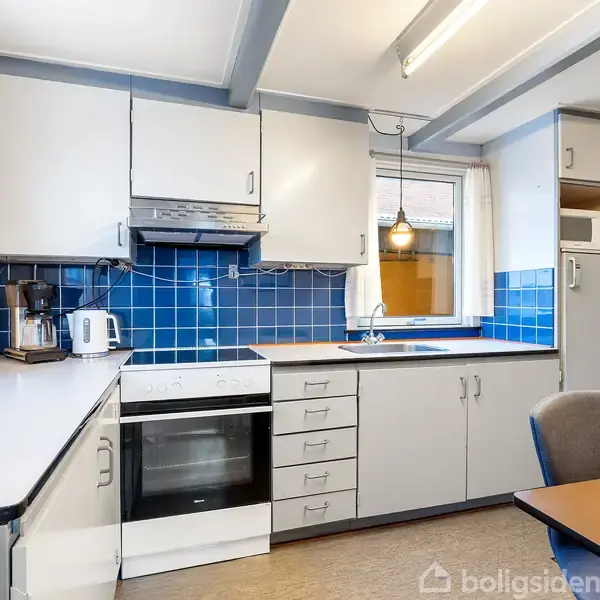 Køkken med beige skabe og blå fliser, indeholder komfur, kaffemaskine og elkedel. Et bord med stol er placeret nær vinduet, der giver naturligt lys.