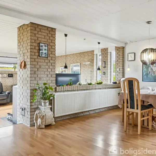 Et åbent opholdsrum med træmøbler står rundt om et spisebord dækket med dug. I baggrunden ses en lænestol og en sofa nær en murstensvæg med planter.