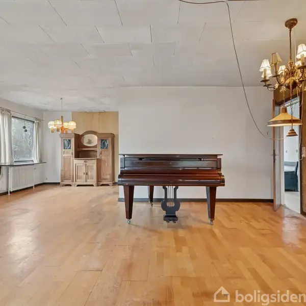 Et flygel står i centrum af en stor, tom stue med trægulve. To lysekroner hænger fra loftet, og i baggrunden ses et skab ved siden af et vindue.