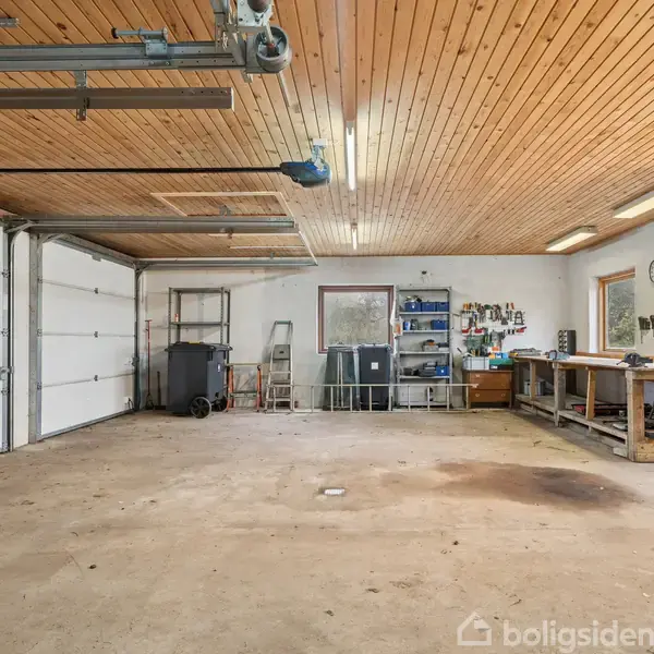 Rummelig garage med to elektriske porte, værktøjsborde til højre og hylder bagerst, omgivet af træloft og lyse vægge.