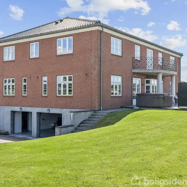 En stor murstensvilla står på en grøn græsplæne. Bygningen har to etager, vinduer, dobbelt garageport og en rummelig terrasse på bagsiden under en blå himmel.