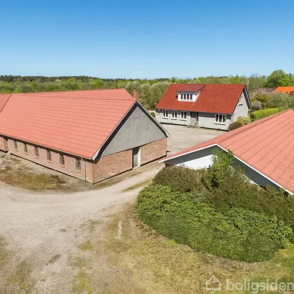 Tre bygninger står roligt i en åbning omgivet af frodig grøn vegetation, med røde tegltage og grusbelægning mellem dem, under en skyfri blå himmel.