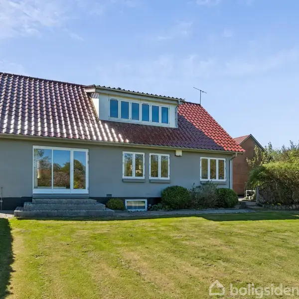 Hus med rødt tegltag står i en velplejet have med græsplæne og hække. Solen skinner på en blå skyfri himmel.