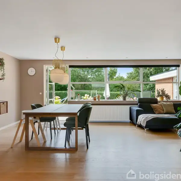 En moderne stue med et spisebord i træ, omgivet af stole, står ved et stort vindue med udsigt til en have. Der er en sort sofa ved siden af, og hylder med dekorationer på væggen.