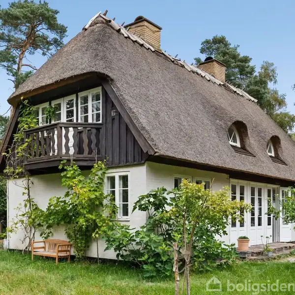 Hus med stråtag ligger i en frodig have omgivet af træer. Husets facade er hvid med mørkt træværk, og en lille balkon ses øverst.