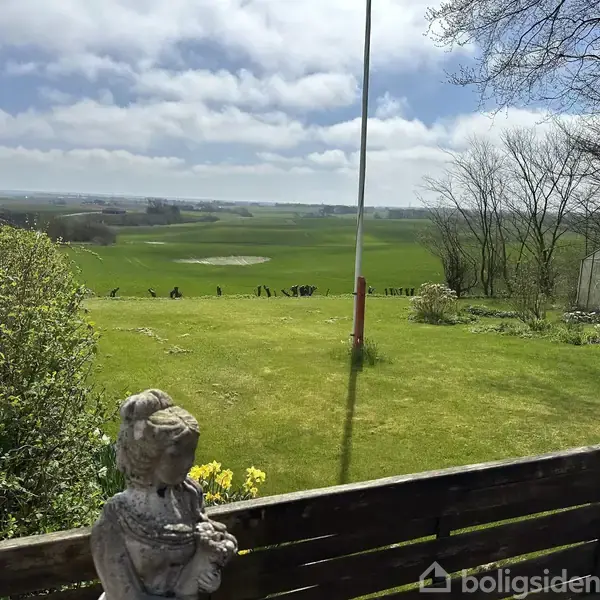 Stenstatue står stille på altanens rækværk. Grønt græs breder sig til horisonten, med spredte træer og en flagstang i det åbne landskabs baggrund. Ingen tekst fundet.
