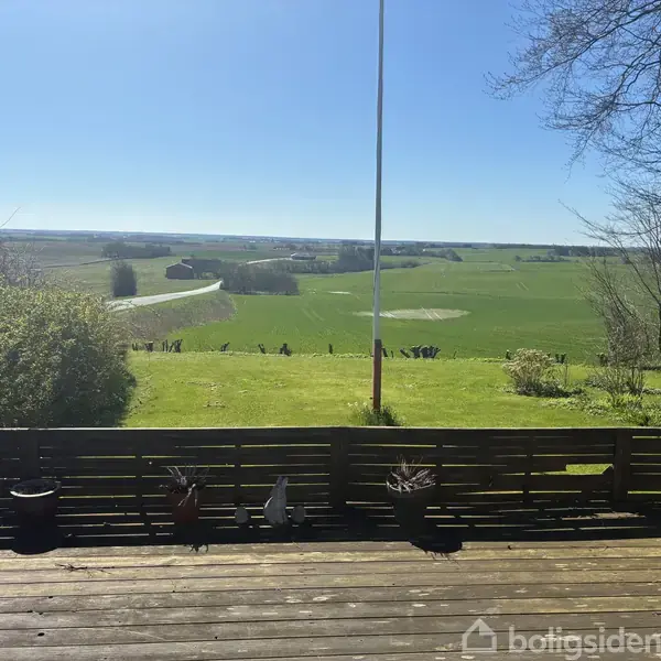 Terrasse med træplankegulv og potteplanter i forgrunden. Græsplæne og flagstang leder til åbne, grønne marker, hvor en vej snor sig gennem landskabet under en klar blå himmel.