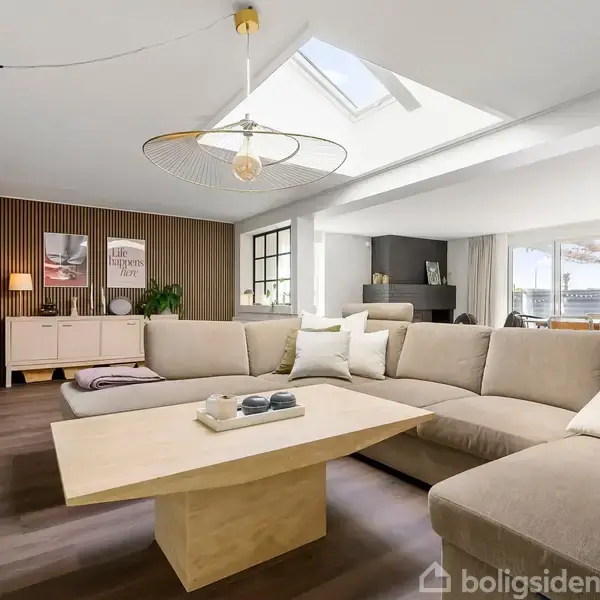 Stor beige sofa står placeret i en rummelig stue med trægulv og træpanelvæg. Et træbord er centralt placeret foran sofaen. Skarpt loftslys og dekor fra sidebord kompletterer indretningen.