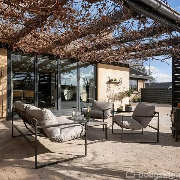 Terrassemøbler med bløde puder står under et pergola overdækket med grene foran en moderne bygning med store vinduer. Terrassefliser og dekorative planter skaber en hyggelig udendørs atmosfære.