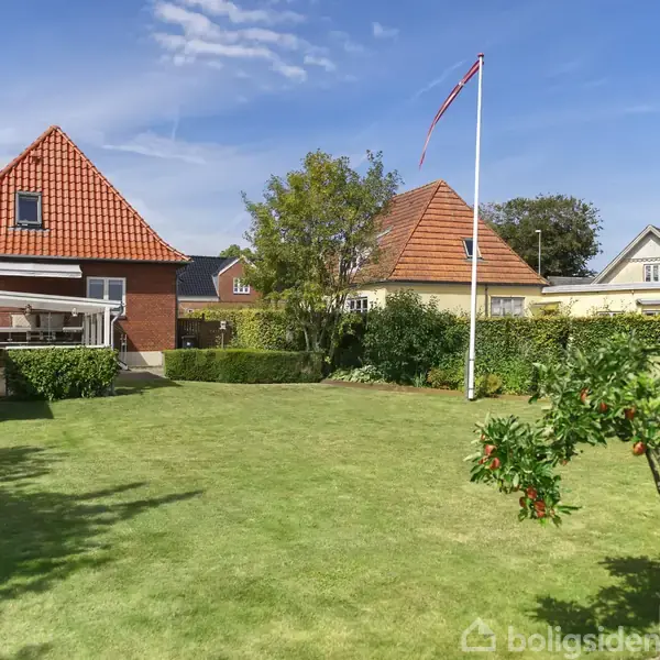 Et græsareal med et æbletræ i forgrunden og to huse med røde tage i baggrunden. En flagstang står til højre, omgivet af hække og træer under en klar himmel.