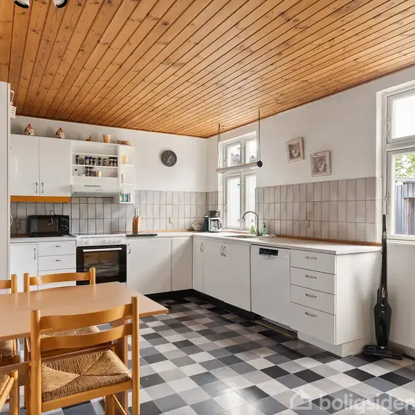 Køkken med bord, stole og hvidt køkkenudstyr. Skakternet gulv og træloft. Rummets lysindfald fra vinduerne skaber en lys og varm atmosfære. Peberkværne på bordet.