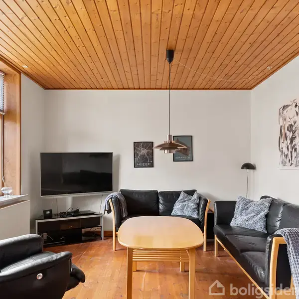 Stue med sort lædersofa og stol, rundt træbord og væghængt fjernsyn. Træbeklædt loft, store vinduer til venstre, lamper og dekorationer på væggene skaber en hyggelig atmosfære.