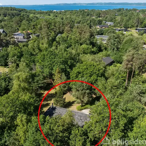 Et hus omgivet af tæt skov ses fra oven, markeret med en rød cirkel. I baggrunden anes flere huse og en havudsigt.