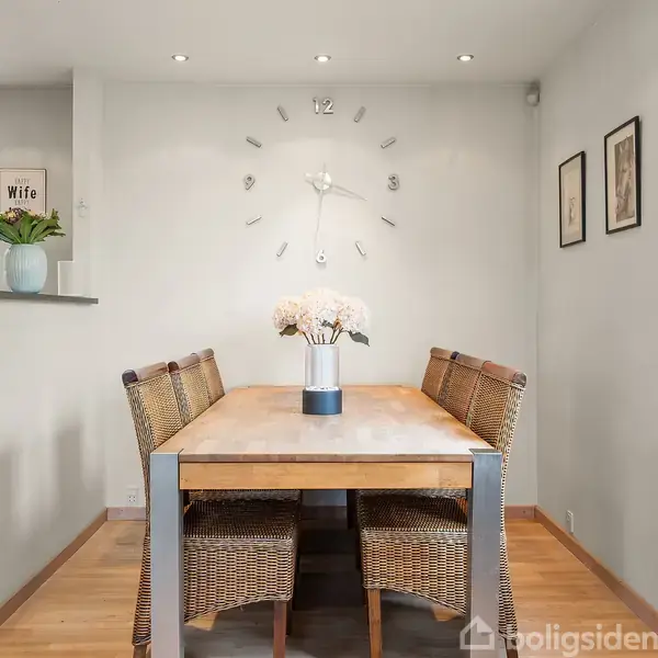 Et træbord med fire fletstole omkring står i en spisestue. En vase med blomster er placeret på bordet. Der er en vægur på væggen, og billedrammer hænger til højre. Tekst på en hylde: "Wife."