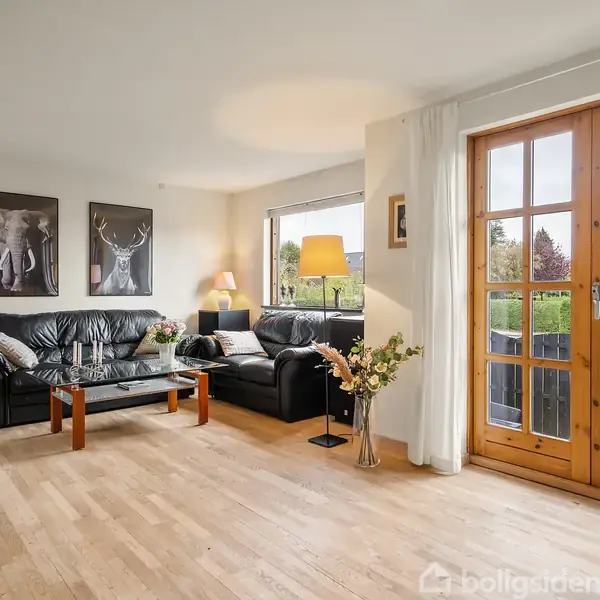 En stue med sort lædersofa, sofabord og lampe, placeret foran store vinduer med udsigt til en have. Trægulv og kunst på væggene komplementerer rummets lyse indretning.