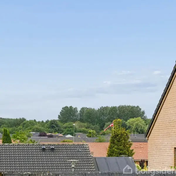 Teglstenstag på huse i forgrunden. I baggrunden står en dansk flagstang ved grønne træer, under en klar blå himmel.