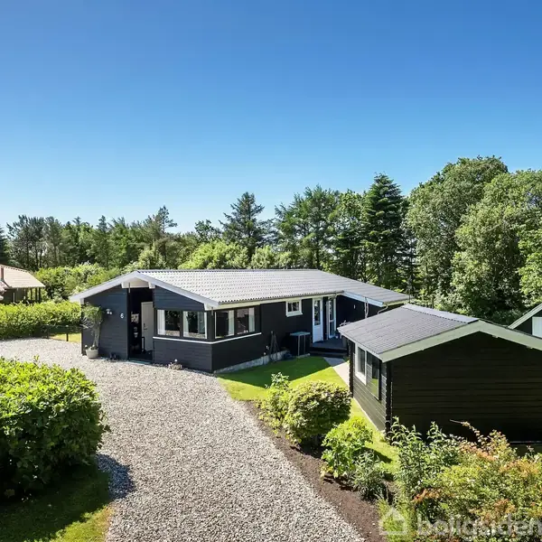 Sort hus med tilhørende skur, omgivet af grønne buske og træer, står på en grusvej i et landligt område. Tætklippet græsplæne omkranser ejendommen under en blå himmel.