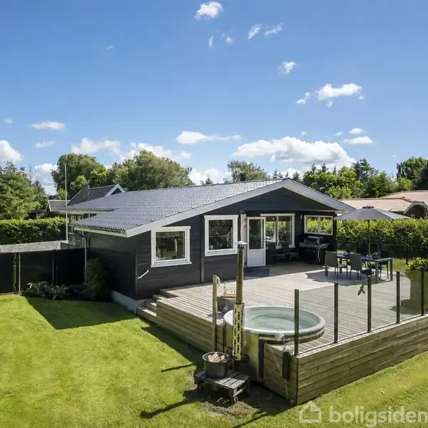 Et moderne hus med hvide vinduer står stille. Det har en stor træterrasse med en rund jacuzzi og glasrækværk. Græsplæne og grønne træer omkranser området under en blå himmel.
