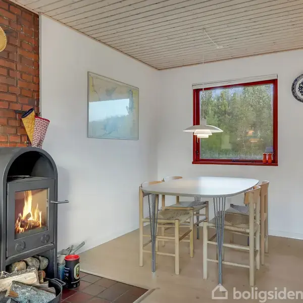 Brændeovn opvarmer hjørne af spisestue; hvidt bord med fire stole; rød indrammet vindue, hvid væg med hængende tallerken; murstensvæg ved siden af. Ingen synlig tekst.