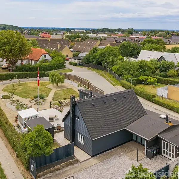 Sort hus med flagstang og flag foran. Omkringliggende haverum med hegn, græs og buske. Flere huse i et villakvarter ses i baggrunden.