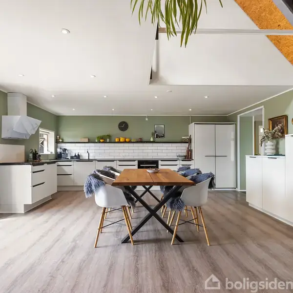 Spisebord i træ med stole, dækket med blå tæpper, står centralt i rummeligt, moderne køkken med grønne vægge og trælamelgulv, omgivet af hvide skabe og hvidevarer.