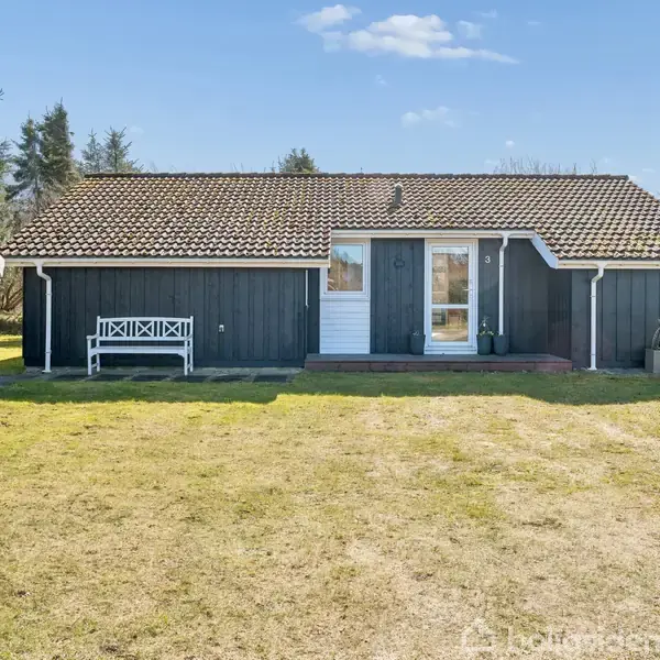 Et mørkt træhus står stille på en græsplæne, med en hvid bænk til venstre. Ved siden af ses en lille sort bygning, omgivet af træer under en blå himmel.
