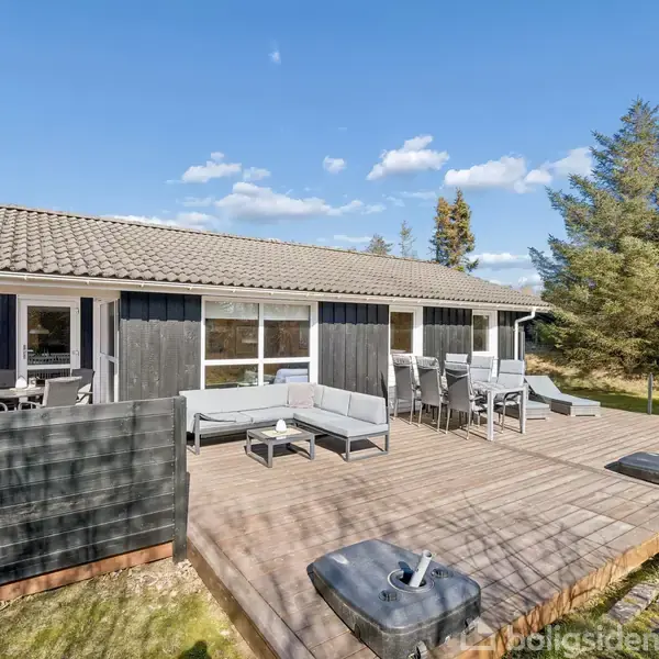 Et sort-hvidt sommerhus står stille på en stor træterrasse, der er indrettet med havemøbler. Omgivet af træer og blå himmel, skaber det en rolig atmosfære.