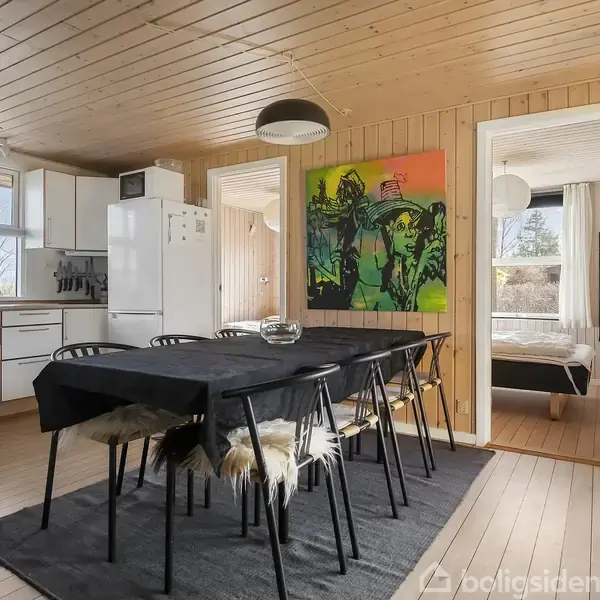 Spisebord med sorte stole står i et træbeklædt rum ved siden af et åbent køkken. Et farverigt maleri hænger på væggen; en dør fører til et tilstødende værelse med seng.