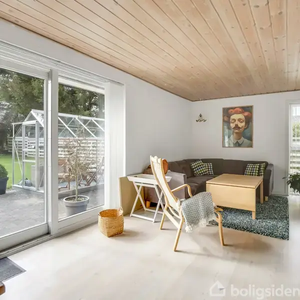 Stue med sofa og træbord, udsigt til have med drivhus gennem store glasdøre. Lyse trægulve og træloft, dekoreret med farverigt vægkunst, minimalistisk indretning.
