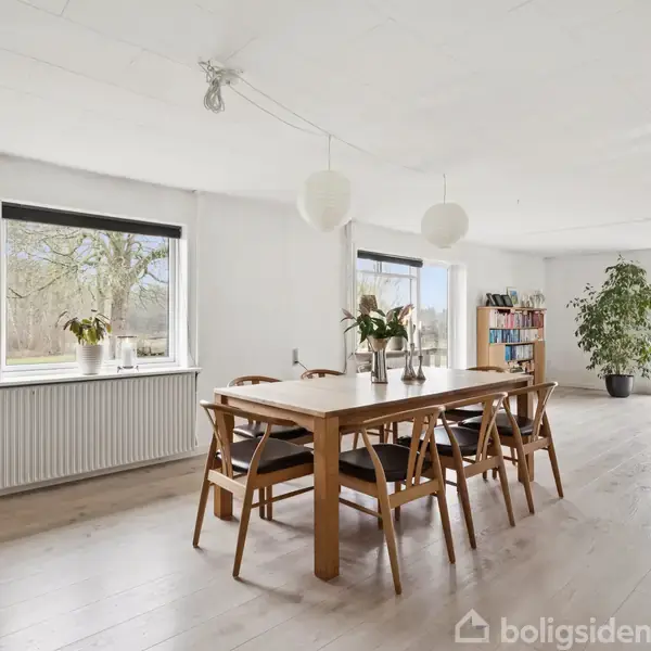Spisebord af træ med stole omkring, prydet med planter i vaser. Rummet er lyst, med store vinduer og indrammes af grønne planter. Reoler i baggrunden.