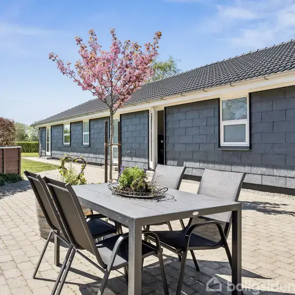 Terrassemøbler står på en flisebelagt gårdsplads ved et moderne hus med grå facade. Et blomstrende træ giver farve, og hække omkranser området i et solrigt landsbymiljø.