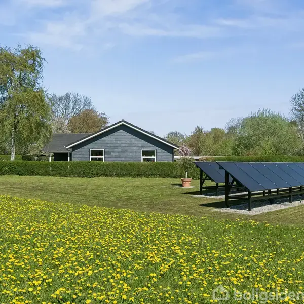 Grå bygning står i en solrig have med gule blomster. Solpaneler er placeret på græsplænen. Omgivet af træer og klar, blå himmel.