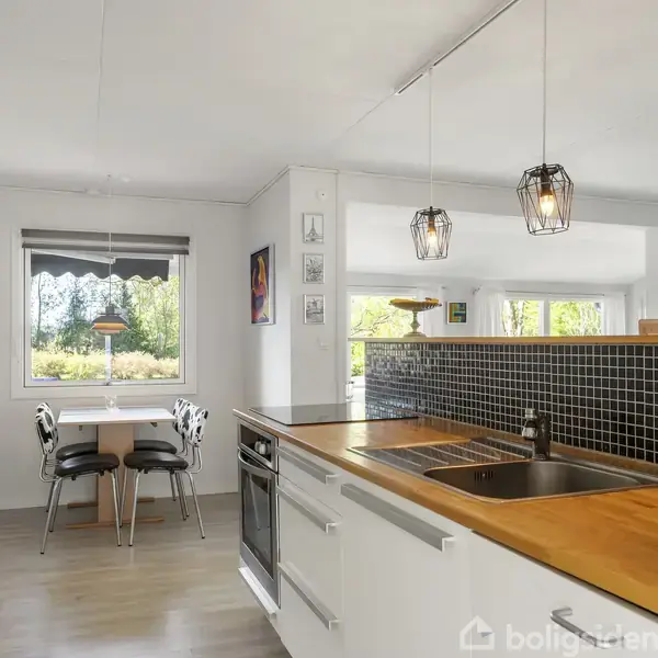Køkken med hvidt interiør og træbordplade. Der hænger moderne lamper over køkkenbordet. I baggrunden er der et spisebord ved et vindue, der viser en grøn udsigt.