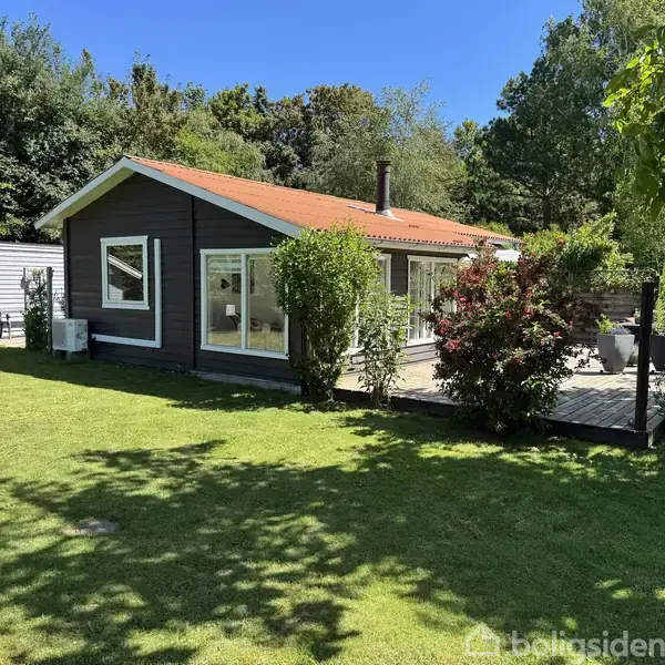 Et mørkegråt sommerhus med rødt tegltag står i solen omgivet af en velplejet have. En træterrasse med havemøbler ses ved huset, omgivet af grønne buske og træer.