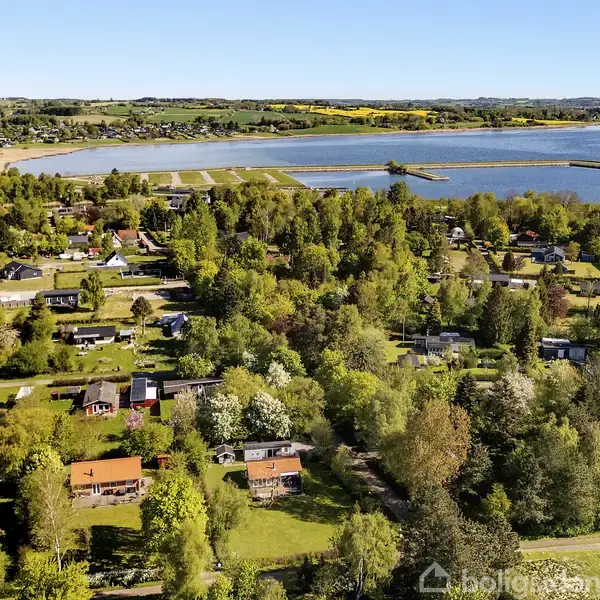Et landområde med spredte huse omringet af tætte grønne træer, der strækker sig mod en bred sø. En landevej snor sig gennem landskabet. Boligsiden vandmærke nederst til højre.