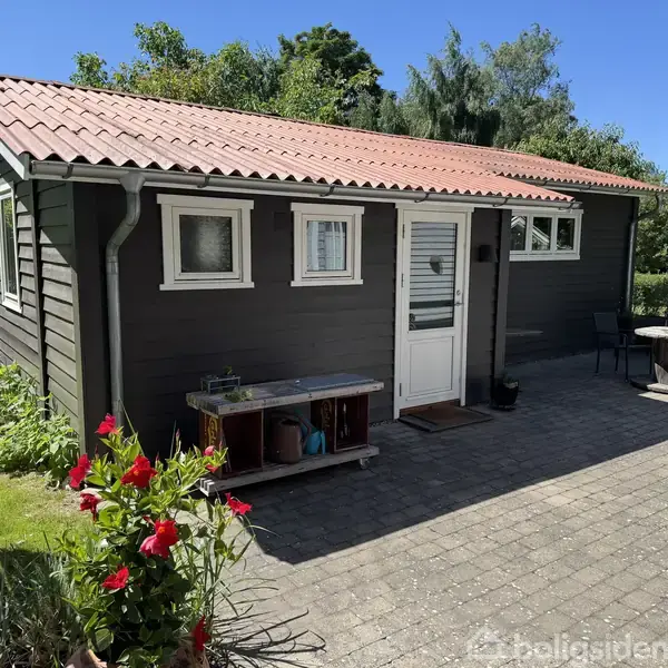 Lille, træbeklædt hus står på en flisebelagt terrasse med rødt tag. Blomster vokser foran huset, mens havemøbler og planter omgiver det i en grøn have.
