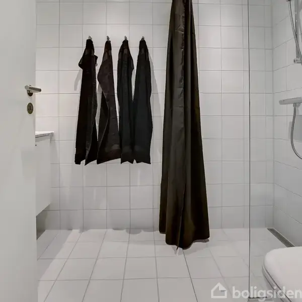 Badeværelse med hvide fliser. En sort bruserforhæng hænger i midten. På væggen hænger mørke håndklæder, og et brusehoved er monteret til højre.