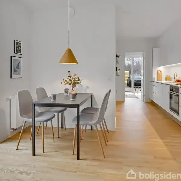 Et moderne spisebord med fire stole står i et lyst køkken-alrum. En gul lampe hænger over bordet, og en vase med blomster pynter bordet. En stor vinduesrude lader naturligt lys ind.