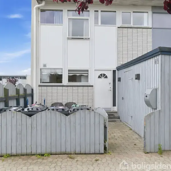 Rækkehus med hvide og grå facader står roligt ved siden af et skur. Affaldsbeholdere er indhegnet foran huset. Bilen holder på græsplæne i forstadsområde.