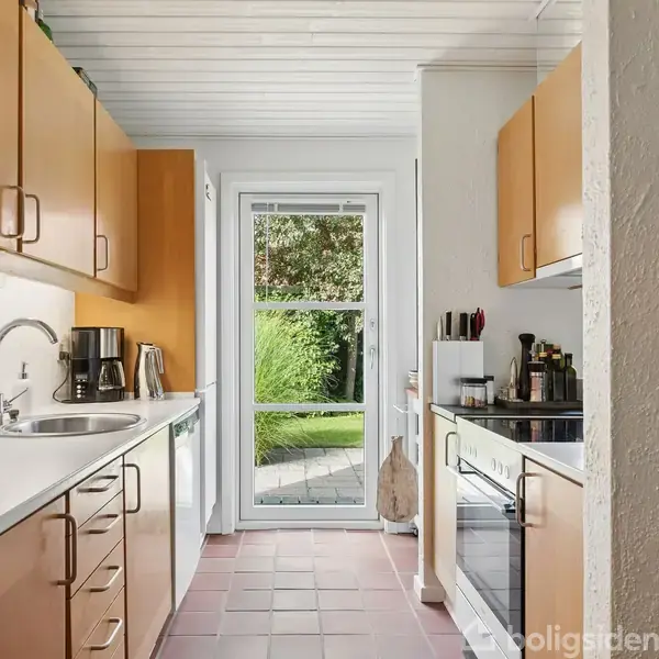 Køkken med træskabe og hvid bordplade, hvor der står en kaffemaskine. En dør med glas fører ud til en have med græs og træer.