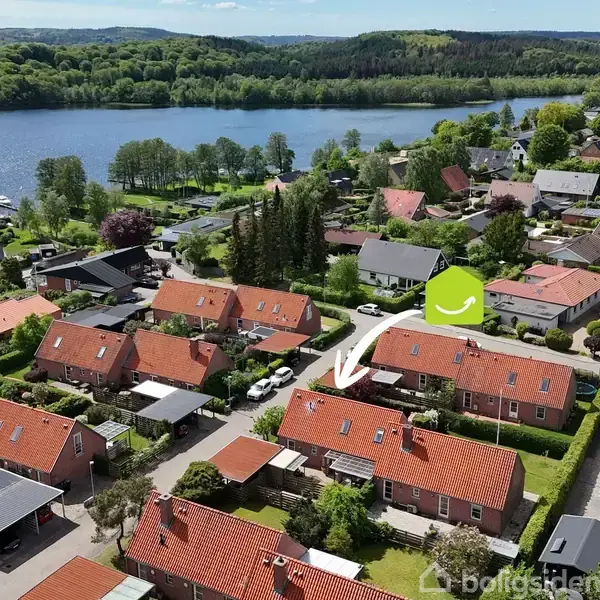 Rækkehuse med røde tage ligger tæt på hinanden. En hvid pil peger på et hus, omgivet af grønne træer. En sø og skov er i baggrunden.