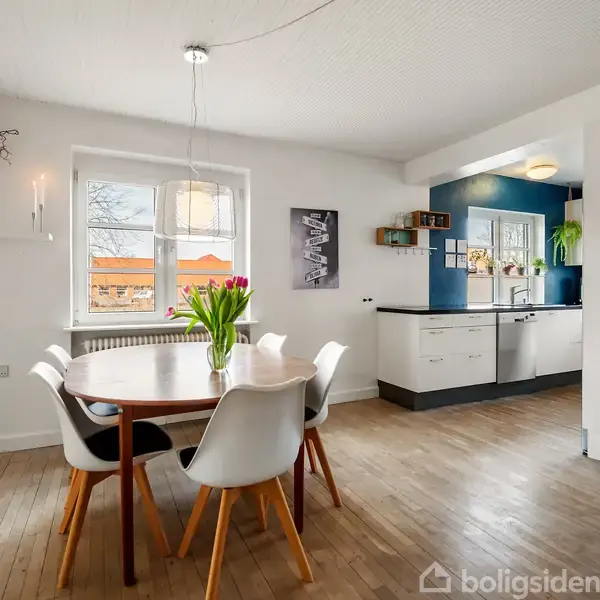 Et rundt spisebord med hvide stole står i et lyst køkken-alrum. Bordet har en vase med tulipaner. I baggrunden ses køkkenskabe, en åben hylde og et vindue.