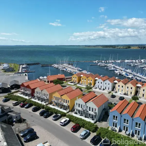 Farverige rækkehuse med røde tage står i flere rækker nær marinaen, hvor lystbåde ligger til kaj. Havet strækker sig ud i horisonten, og himlen er klar med skyer.