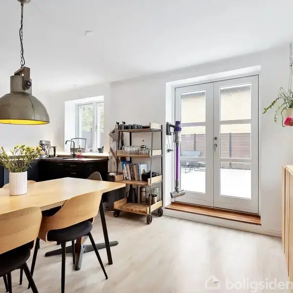 Spisebord med stole står i et moderne køkken. Køkkenet har sorte apparater, trægulv og glasdør til en terrasse. En vase med blomster pryder bordet, omgivet af naturligt lys.