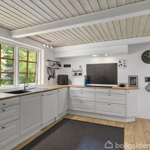 Hvidt køkken med træbordplader, indbyggede skabe og vask, beliggende i et rum med træloftsbjælker og et stort vindue med udsigt til frodige træer. Væghængt ur og køkkenredskaber.