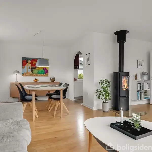 Brændeovn står i stue, spreder varme, omgivet af moderne indretning med lyse vægge, sofabord, sofa og spisebord med stole. Kunstmaleri og planter tilføjer farve og liv.