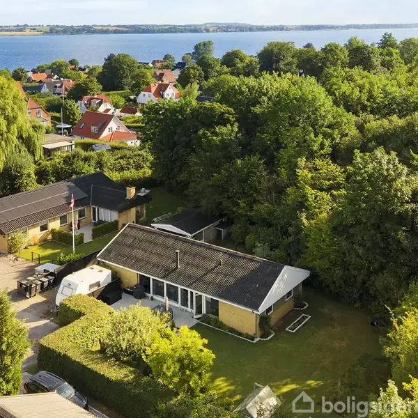 Et hus står i en grøn have omgivet af træer, med udsigt til en sø i baggrunden. Flere huse kan ses nær vandet i den fredelige landsby.