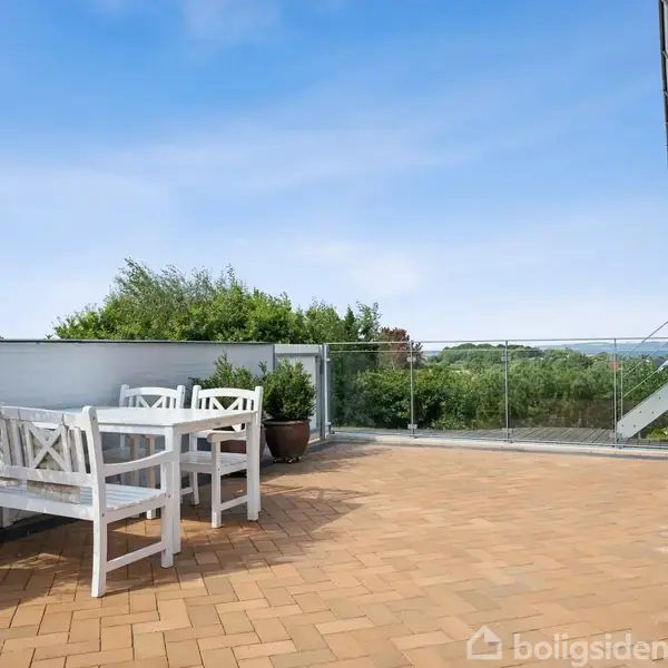 Hvide havemøbler står på en flisebelagt terrasse med glasrækværk. I baggrunden er der grønne planter og udsigt over landskab og himmel.