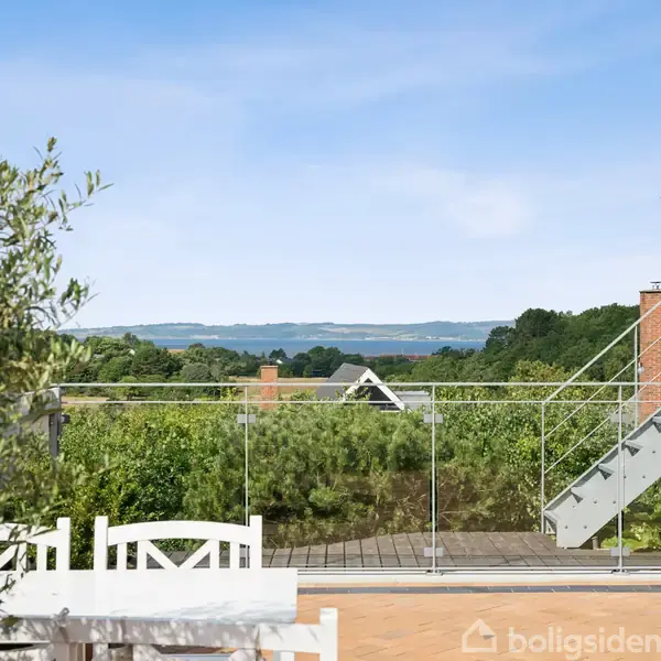 En terrasse med glasrækværk tilbyder udsigt over frodige grønne træer og buske, med en sø og landlige bakker i baggrunden under en klar blå himmel.