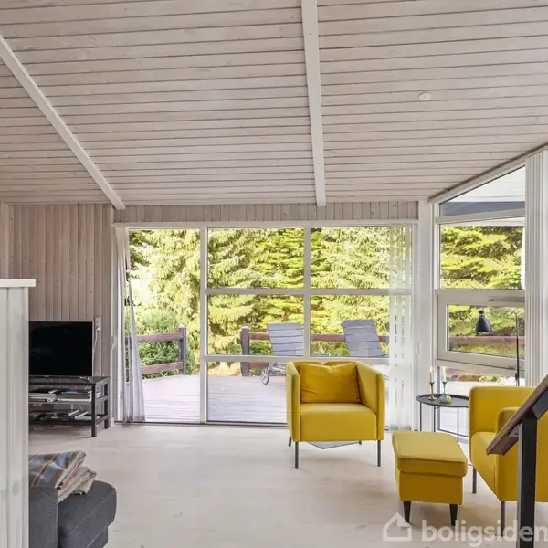 Gule lænestole står i et lyst, moderne stueområde med store vinduer, der åbner op til en terrasse. Udenfor ses grønne træer. En fladskærm står i hjørnet.